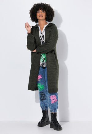 Donna dai capelli ricci che indossa un lungo cardigan verde scuro, una maglietta grafica bianca, pantaloni jogger stampati e colorati, e stivali neri con lacci, in piedi contro un muro bianco.