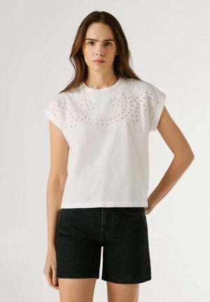 Mujer con camiseta blanca de manga corta con bordado floral de ojales y shorts de mezclilla negros, de pie con una mano en la cadera contra un fondo liso.