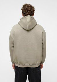 Überdimensionierter Kapuzenpullover in hellbeige, mit strukturiertem Stoff, gerippten Bündchen und Saum. Getragen mit dunkelgrauen Hosen.