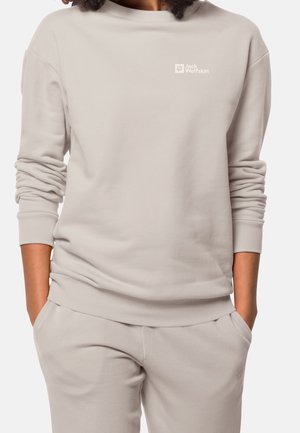 Osoba ubrana w jasnobeżową bluzę Jack Wolfskin i dopasowane spodnie z rękami w kieszeniach, widoczna od szyi do bioder.