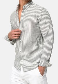 Homme portant une chemise à rayures verticales grises et blanches, manches retroussées, et un pantalon blanc, ajustant sa chemise près de la poitrine.