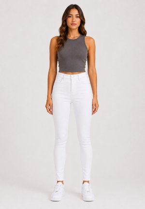 Vrouw staand, dragend een donkergrijs mouwloos crop top, witte high-waisted skinny jeans en witte sneakers tegen een effen achtergrond.