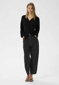 Melna pogu jaciņa ar V veida kakla izgriezumu, saskaņota ar augsto vidukli un brīvā stila melnajām džinsām. Modelis valkā plakanas melnas sandales.