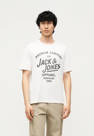 Młody mężczyzna o czarnych kręconych włosach, ubrany w białą koszulkę z logo "Jack & Jones" i beżowe krótkie spodnie cargo na jednolitym tle.