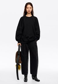 Sudadera negra con detalles de tachuelas en los hombros, combinada con pantalones negros holgados y un bolso marrón con un tassel amarillo.