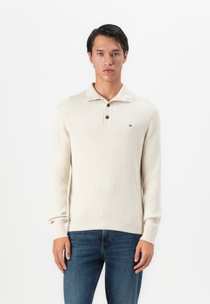 Mann in einem cremefarbenen, gerippten Strickpullover mit einem geknöpften Kragen und blauen Jeans, der vor einem weißen Hintergrund steht.