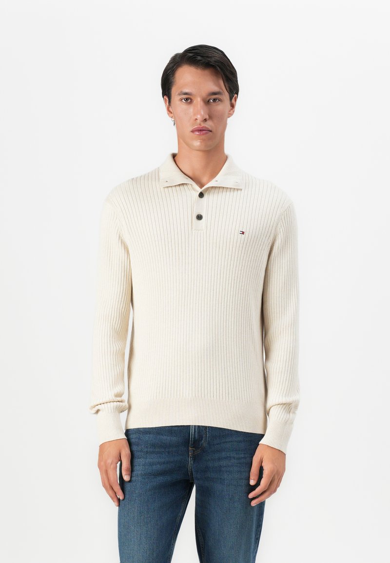 Homme portant un pull en maille côtelée crème avec un col boutonné et un jean bleu, se tenant contre un fond blanc.
