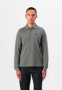 Langarm-Poloshirt aus olivgrünem Stoff, mit Kragen und einem kleinen Logopatch auf der linken Brust. Texturierte Oberflächendetails sind sichtbar.