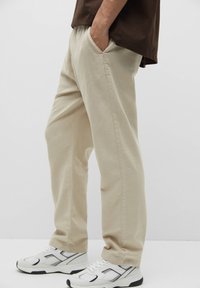 Persona che indossa pantaloni casual beige e sneakers bianchi, con una mano in tasca, una camicia marrone visibile dalla vita in giù.
