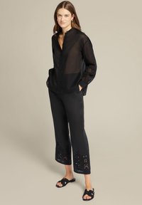 Blusa nera trasparente con colletto alto, dettaglio di bottoni sulla patta e maniche lunghe. Pantaloni neri con ricami alla caviglia. Sandali neri.
