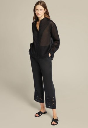 Blusa nera trasparente con colletto alto, dettaglio di bottoni sulla patta e maniche lunghe. Pantaloni neri con ricami alla caviglia. Sandali neri.
