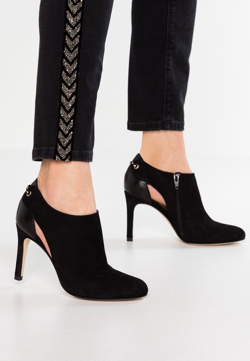 Bottines à talons hauts en suède noir avec des côtés découpés, dotées d'une fermeture éclair et d'accents dorés. Couleur noire uni avec une texture lisse.