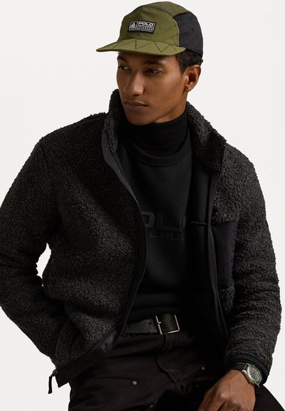 Črna flis jakna z teksturirano tkanino, z zadrgo spredaj, v kombinaciji s črnim puloverjem s stojalom in z zeleni/črnim klobukom z logotipom. Vidna črna pas in ura.