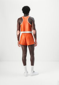 Top sportivo in mesh arancione con pantaloni corti e rifiniture bianche. Il modello indossa calze e scarpe bianche, mostrando la pelle texturizzata su braccia e gambe.