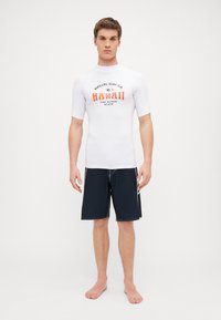 Hvit kortermet surfeskjorte med rød og svart logo-tekst, kombinert med marineblå svømshorts med hvit kontrastsøm.