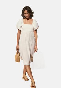 Robe blanche à manches bouffantes avec décolleté carré, corsage cintré, jupe fluide et longueur mi-mollet. Associée à des sandales à lanières marron et un sac en osier.