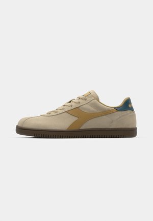 Zapatilla de ante beige con suela de goma marrón, acentos amarillos en el costado y una tapa de talón azul. Diseño con cordones y paneles texturizados.
