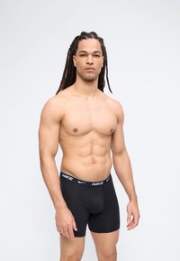 Μυώδης άνδρας με μακριές κοτσίδες dreadlocks που φοράει μαύρα εσώρουχα Nike boxer, στέκεται μπροστά σε απλό λευκό φόντο.