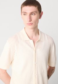 Polo en tricot crème à manches courtes, présentant des rayures verticales, une fermeture à boutons et une texture douce.
