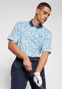 Kortermet polo skjorte i lyseblå med blomstermønster; marineblå krage. Kombinert med mørkeblå bukser, holder en golfkølle, iført en hanske.