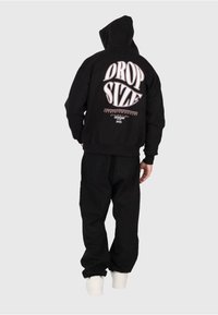 Persona che indossa una felpa nera con il testo "DROP SIZE" sulla schiena, pantaloni neri e sneakers bianche, in piedi contro uno sfondo chiaro e semplice.