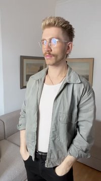 Veste zippée gris clair sur une chemise blanche ajustée, pantalon noir et lunettes transparentes. Cheveux courts en épis, debout avec les mains dans les poches.