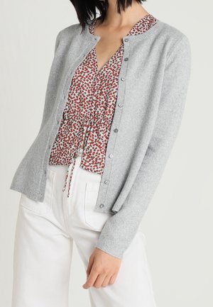 Femme portant un cardigan gris clair boutonné sur un chemisier rouge et blanc à motifs avec un pantalon blanc, debout devant un fond uni.