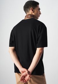 Czarna koszulka z krótkim rękawem o luźnym kroju. Posiada brązowy wzorzysty szalik wokół szyi. Prosty design, gładka tekstura.