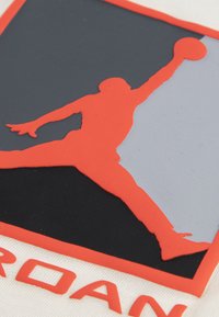 Μαύρο, γκρι και πορτοκαλί λογότυπο που απεικονίζει τη σιλουέτα ενός παίκτη που πηδάει, με υφές από καουτσούκ σε λευκό υφασμάτινο φόντο.