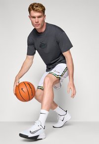 Grå Nike shirt med Lakers-logo, hvide basketballshorts med farverigt mønstret kant, hvide og sorte Nike sneakers, holder en basketball.