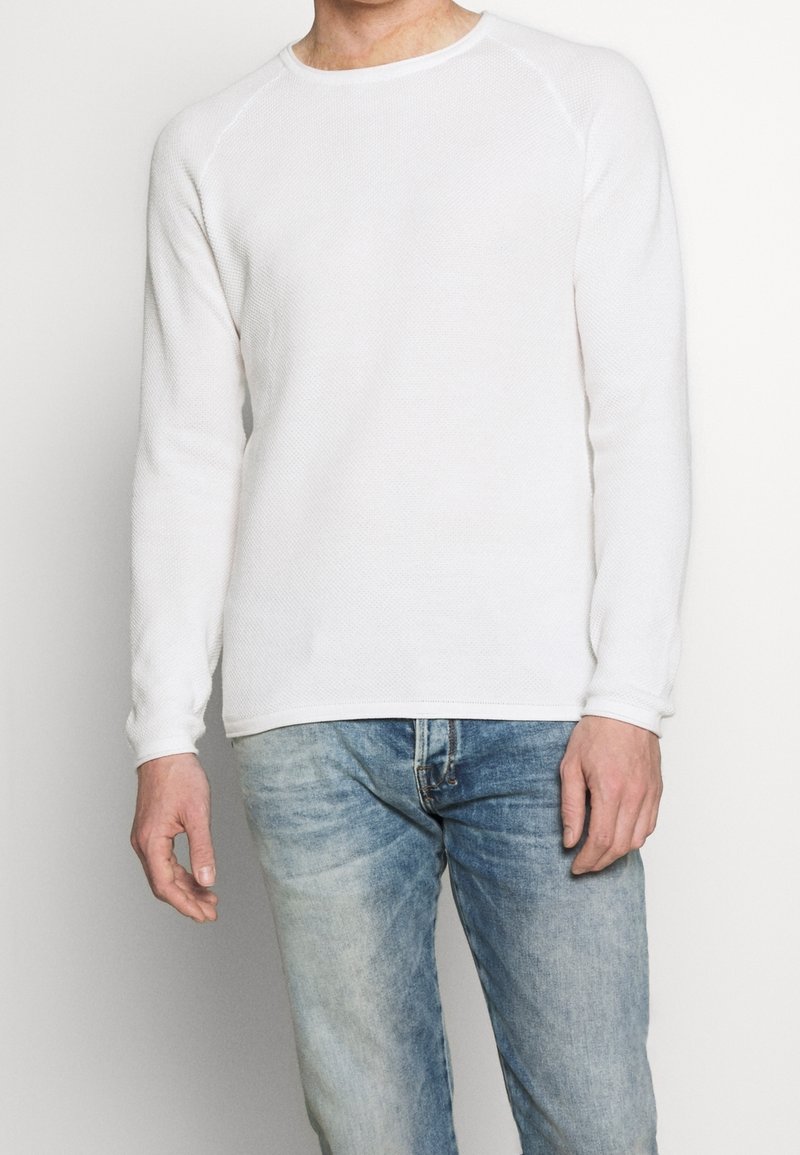 Homme portant une chemise blanche à manches longues texturée et un jean bleu clair délavé, debout devant un fond blanc uni.