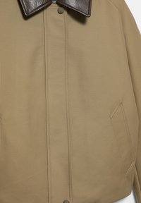 Bež plašč z ovratnikom iz temno rjavega usnja, sprednjimi gumbi na zadrgo, navpičnim šivom in poševnimi stranskimi žepi.