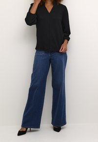 Sort, tekstureret bluse med V-hals og trekvartærmet, parret med brede blå denimjeans og sorte hæle.