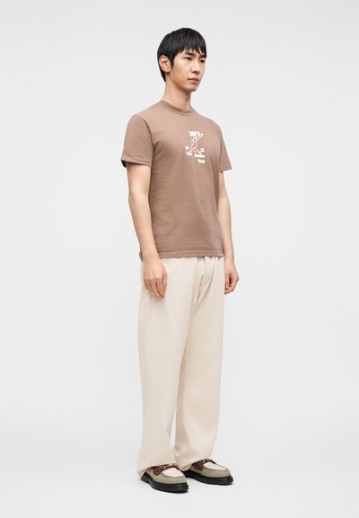 Καφέ κοντομάνικη μπλούζα με γραφικό σχέδιο, σε συνδυασμό με χαλαρής εφαρμογής μπεζ παντελόνια και πολύχρωμα παπούτσια με διακοσμητικά σ τοιχεία και υφή.