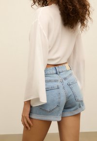 Bílá lehká blůza s zvonovými rukávy, kterou doplňují modré džínové šortky s vysokým pasem a ohrnutými lemy a zadními kapsami.