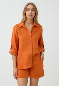 Roestkleurig, textuurshirt met opgerolde mouwen en knoopsluiting, gecombineerd met bijpassende shorts. Beide stukken hebben een relaxed fit.