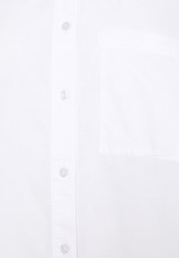 Hvid skjorte med knapper, der har en glat tekstur, seks klare knapper og en lomme på venstre side. Klassisk krave og lange ærmer.