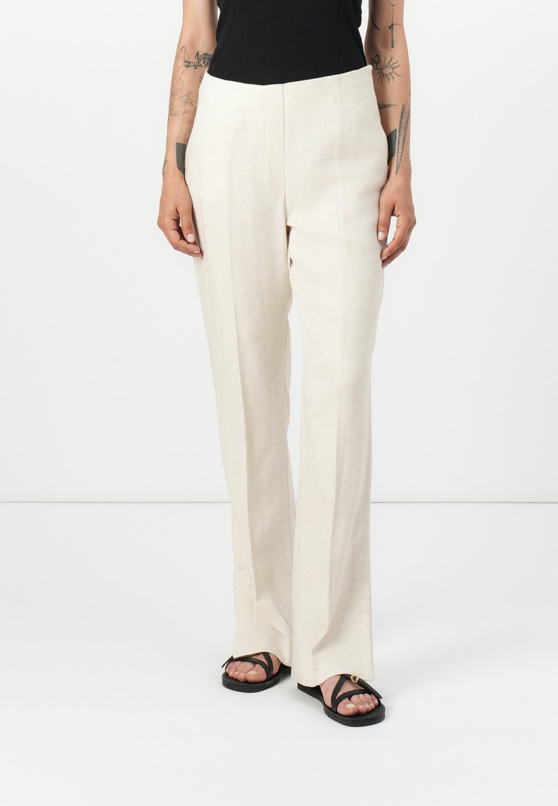 Pantaloni in lino beige con design a gamba dritta, caratterizzati da una texture liscia e sottili cuciture verticali. Abbinati a sandali neri.