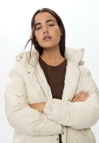 Nafoukaná krémová bunda s vysokým límcem, předním zipem a patentkami. Nositá přes hnědý žebrovaný top, který vystavuje jemné textury.