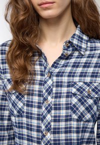 Camicia in flanella a quadri blu navy e bianca con colletto button-down, due tasche sul petto e bottoni dall'aspetto legnoso, caratterizzata da una texture morbida.