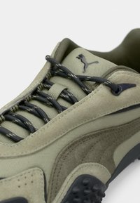 Grüne Wildleder- und Mesh-Sneaker mit dunkelgrauen Schnürsenkeln, strukturierten Akzenten und einem auffälligen Logo in der Nähe des Knöchels. Geschwungenes Design und gemusterte Laufsohle.