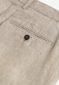 Ljusbeige byxor med en strukturerad tyg, som har en bakficka med en svart knapp och dubbla sömmar längs kanterna.