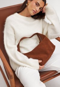 Brązowa torebka z zamszu z zaokrągloną rączką u góry i zamknięciem na zamek, trzymana na białym swetrze i kremowym tle, na drewnianym krześle.