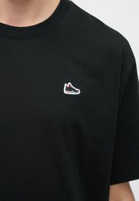 Μαύρο βαμβακερό μπλουζάκι με μικρό κεντητό λογότυπο αθλητικού παπουτσιού σε γκρι, κόκκινο και άσπρο στην αριστερή περιοχή του στήθους. Κοντά μανίκια και λαιμός crew.