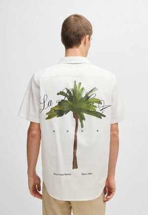 Mężczyzna noszący białą koszulę z krótkim rękawem z zieloną grafiką palmy i czarnym tekstem "La La La," "Hugo," "West Coast Stories," oraz "Since 1993" na plecach.