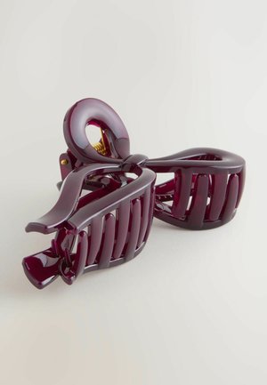 Purple plastic hair clip with a bow design, featuring a glossy finish, curved shape, and interlocking teeth for secure holds. Gold metal hardware visible.