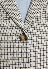 Beige och svart rutigt kavaj med snidade slag, med en enkel rund knapp och en texturerad tygdetalj. Blå tröja synlig under.