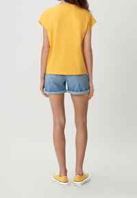 Kobieta widziana z tyłu, ubrana w żółtą koszulkę, podwinięte dżinsowe szorty i żółte trampki, stojąca na jednolitym tle.