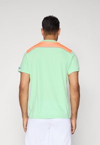 Bărbat văzut din spate, purtând o cămașă scurtă de culoare verde mentă cu un element din plasă portocalie și pantaloni scurți albi, pe un fundal simplu.