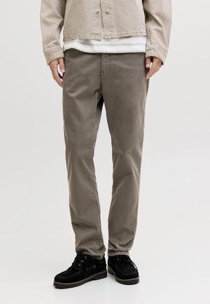 Homme portant un pantalon droit taupe, une veste beige, une chemise blanche superposée et des chaussures noires à lacets, debout sur un fond blanc.